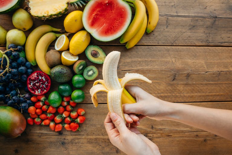 A person peels a banana surrounded by a variety of fresh fruits on a wooden table.