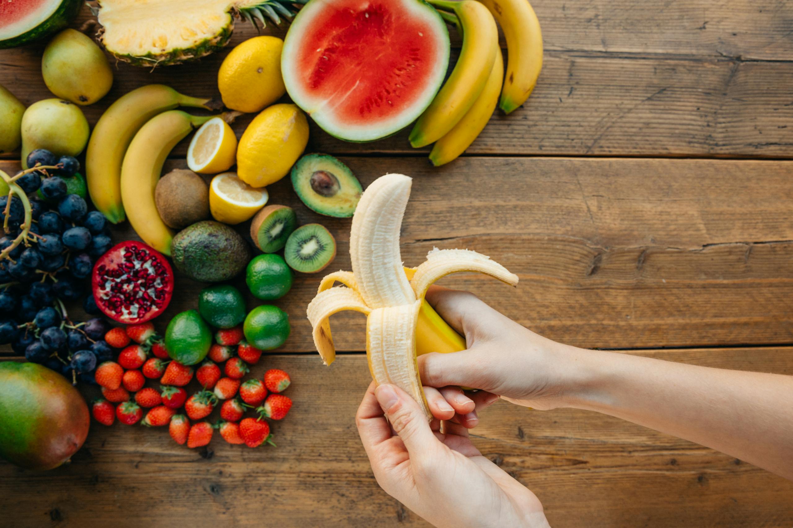 A person peels a banana surrounded by a variety of fresh fruits on a wooden table.