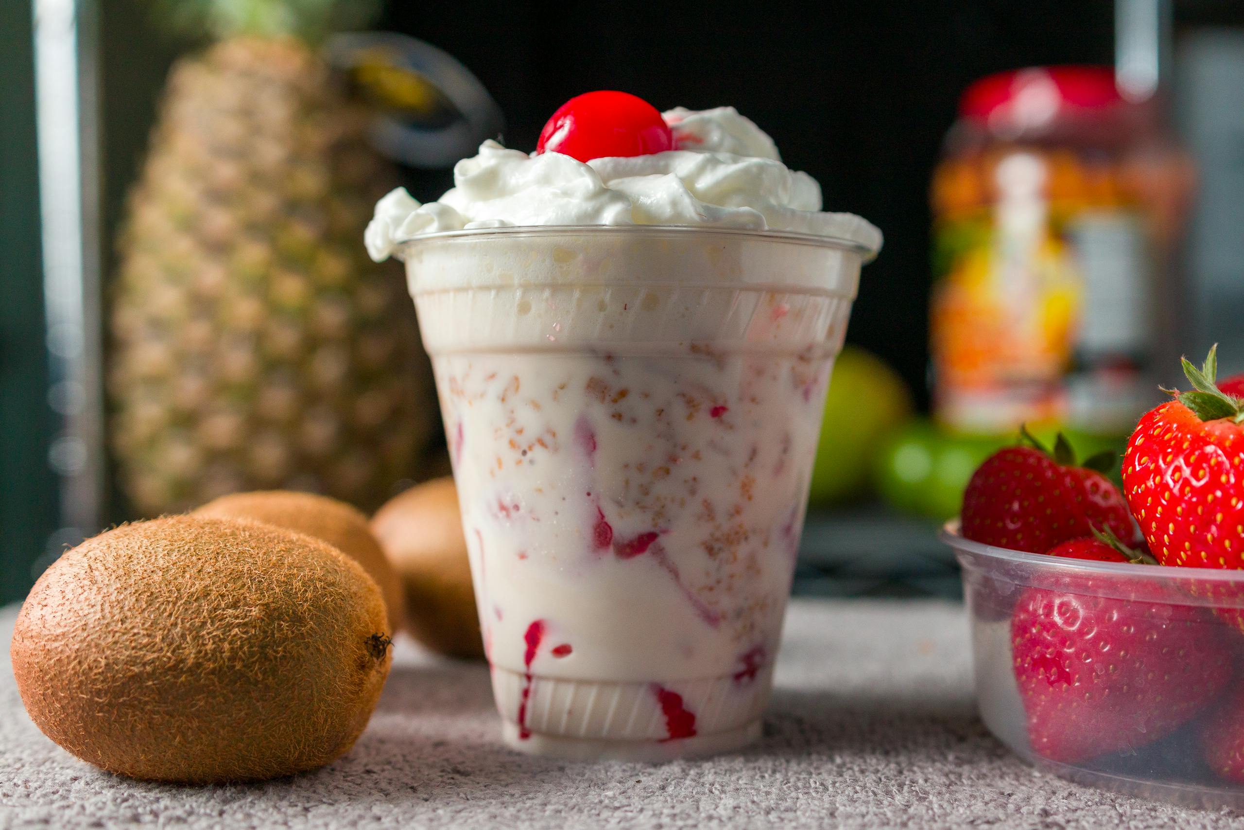 Close-up of a creamy fruit smoothie topped with whipped cream and cherry, surrounded by fresh fruit.